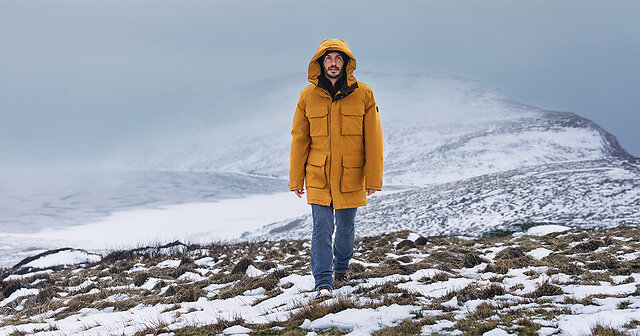 Mann steht in einer senfgelben Winterjacke auf den Bergen im Schnee