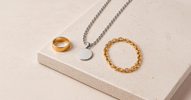 Schmuckstücke auf hellem Untergrund, darunter ein goldener Ring, eine silberne Halskette mit Anhänger und eine goldene Kette